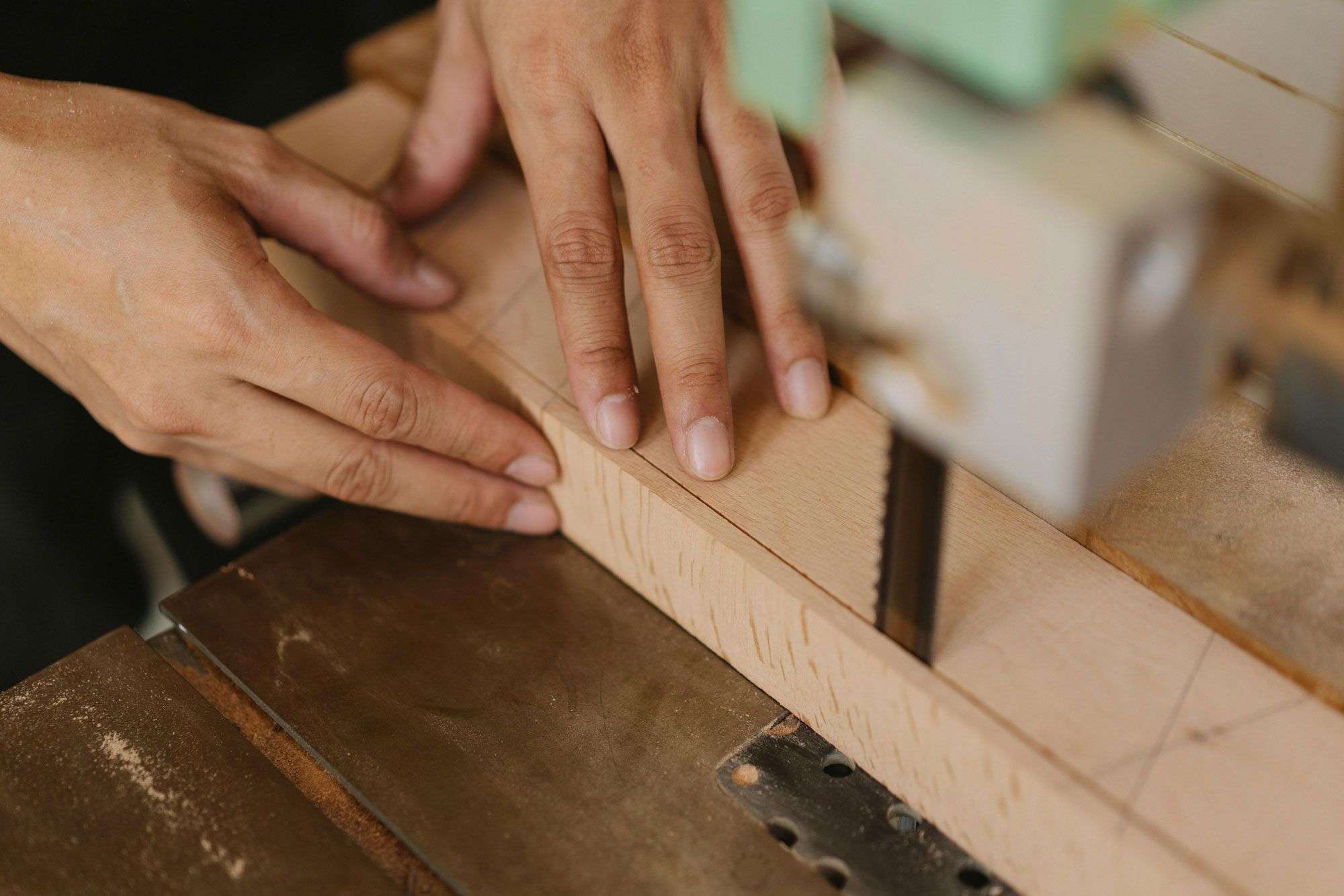Hände einer Person führen präzise ein Holzstück durch eine Bandsäge. Die Szene zeigt handwerkliche Arbeit und den sorgfältigen Umgang mit Holz in einer Werkstatt.