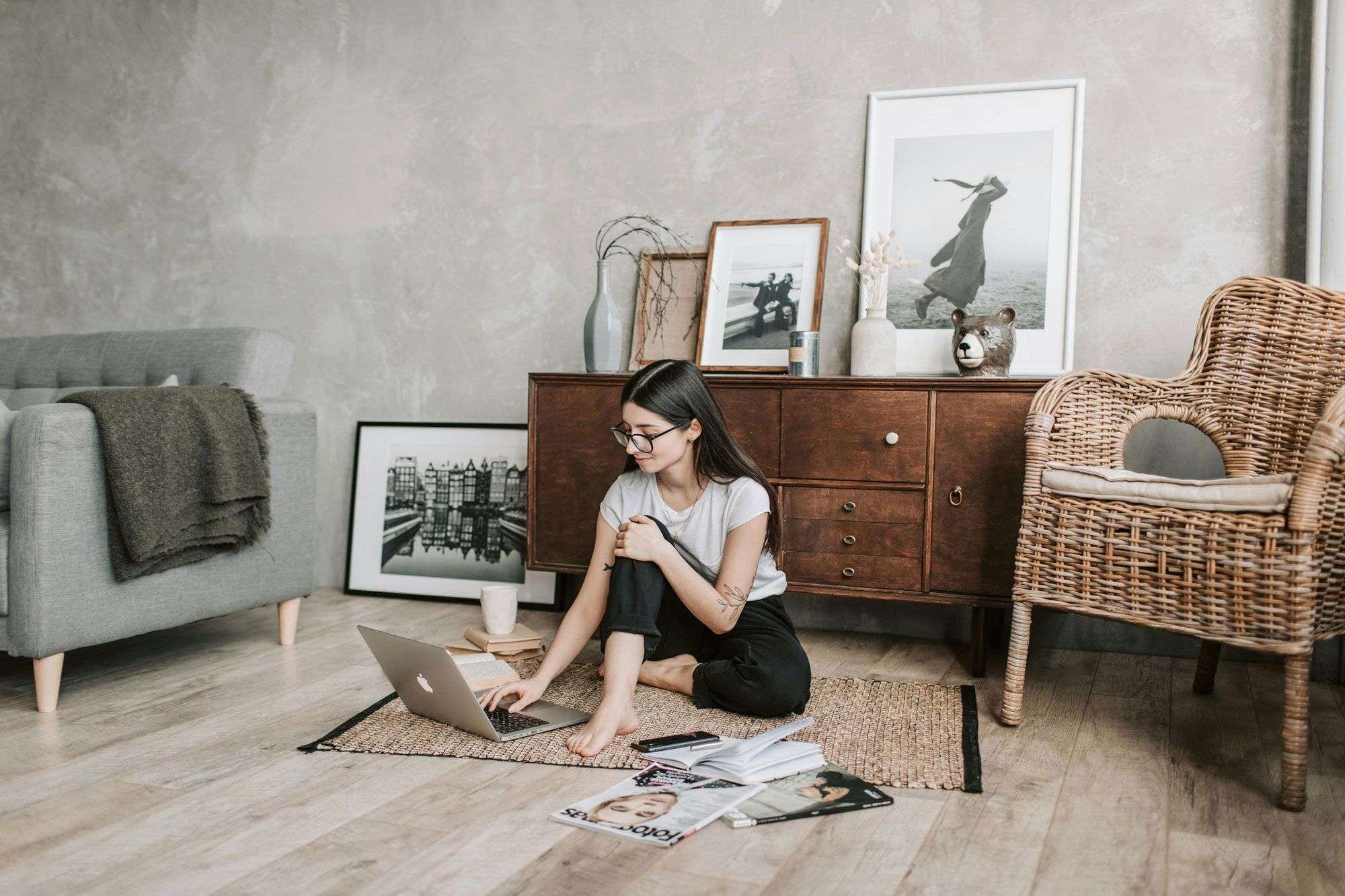 Eine Person sitzt barfuß auf einem Teppichboden und arbeitet an einem Laptop. Um sie herum liegen Zeitschriften, Bücher und eine Tasse. Im Hintergrund steht ein Schrank mit dekorativen Bildern und einem Rattan-Stuhl.