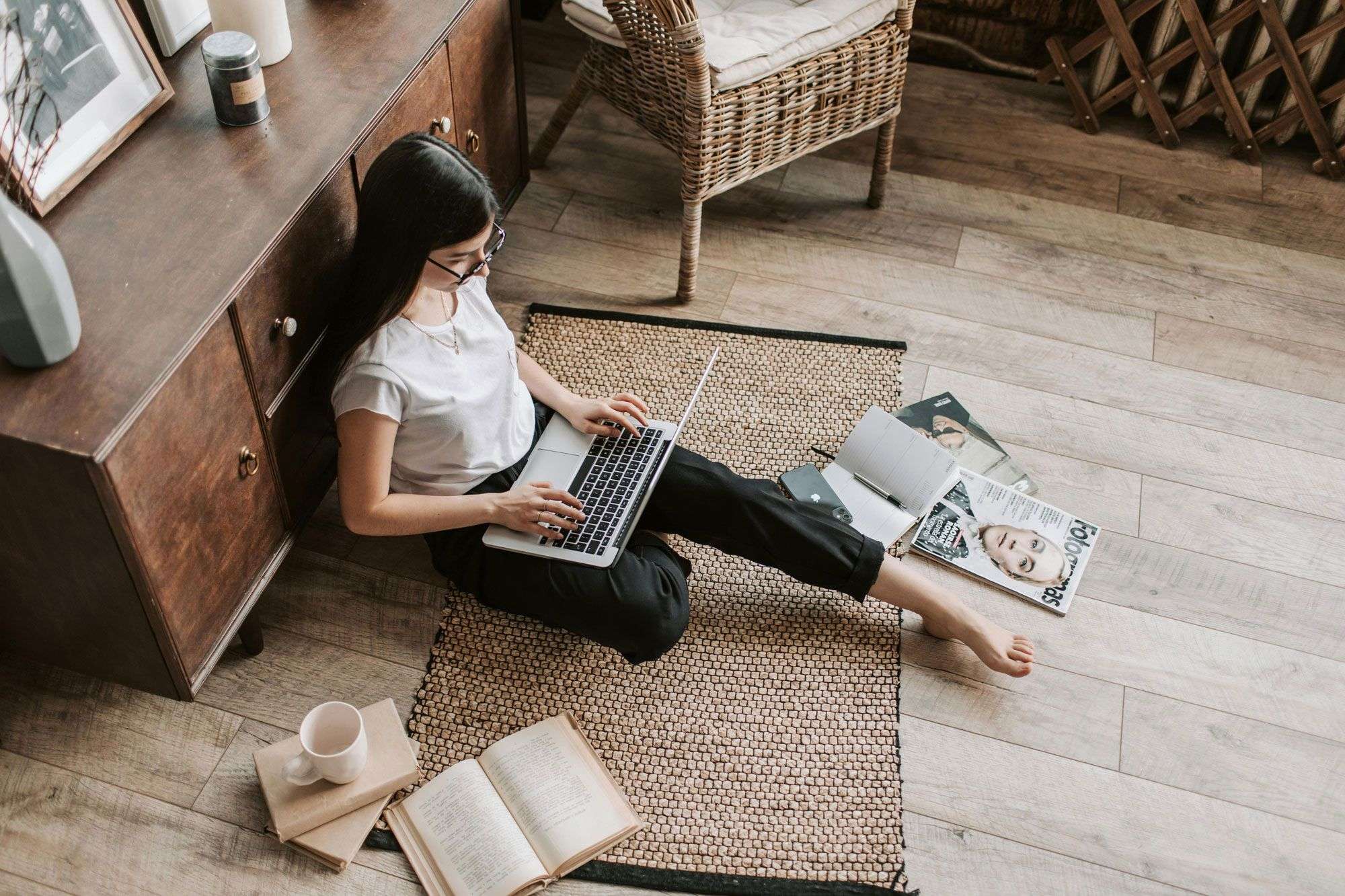 Eine Person sitzt barfuß auf einem Teppichboden, lehnt an einem Schrank und arbeitet an einem Laptop. Um sie herum liegen Bücher, eine Tasse und Zeitschriften verstreut.