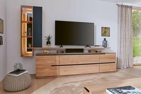 Wohnzimmer mit moderner Wohnwand aus hellem Holz und schwarzen Akzenten. Ein großer Fernseher steht auf der niedrigen Kommode, daneben ein offenes Regal mit integrierter Beleuchtung. Der Raum wirkt hell und freundlich durch die großen Fenster und natürlichen Materialien.