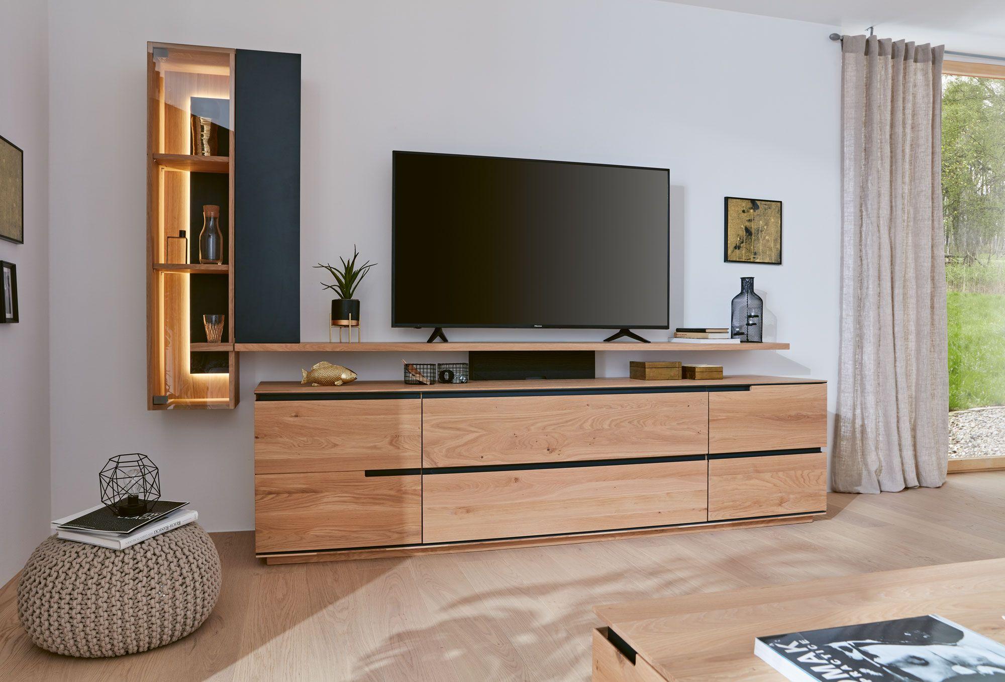 Wohnzimmer mit moderner Wohnwand aus hellem Holz und schwarzen Akzenten. Ein großer Fernseher steht auf der niedrigen Kommode, daneben ein offenes Regal mit integrierter Beleuchtung. Der Raum wirkt hell und freundlich durch die großen Fenster und natürlichen Materialien.