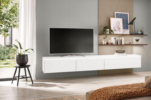 Schwebendes weißes Lowboard mit Fernseher an einer modernen Wand, ergänzt durch Holzdetails, dekorative Regale und Pflanzen, in einem lichtdurchfluteten Raum.