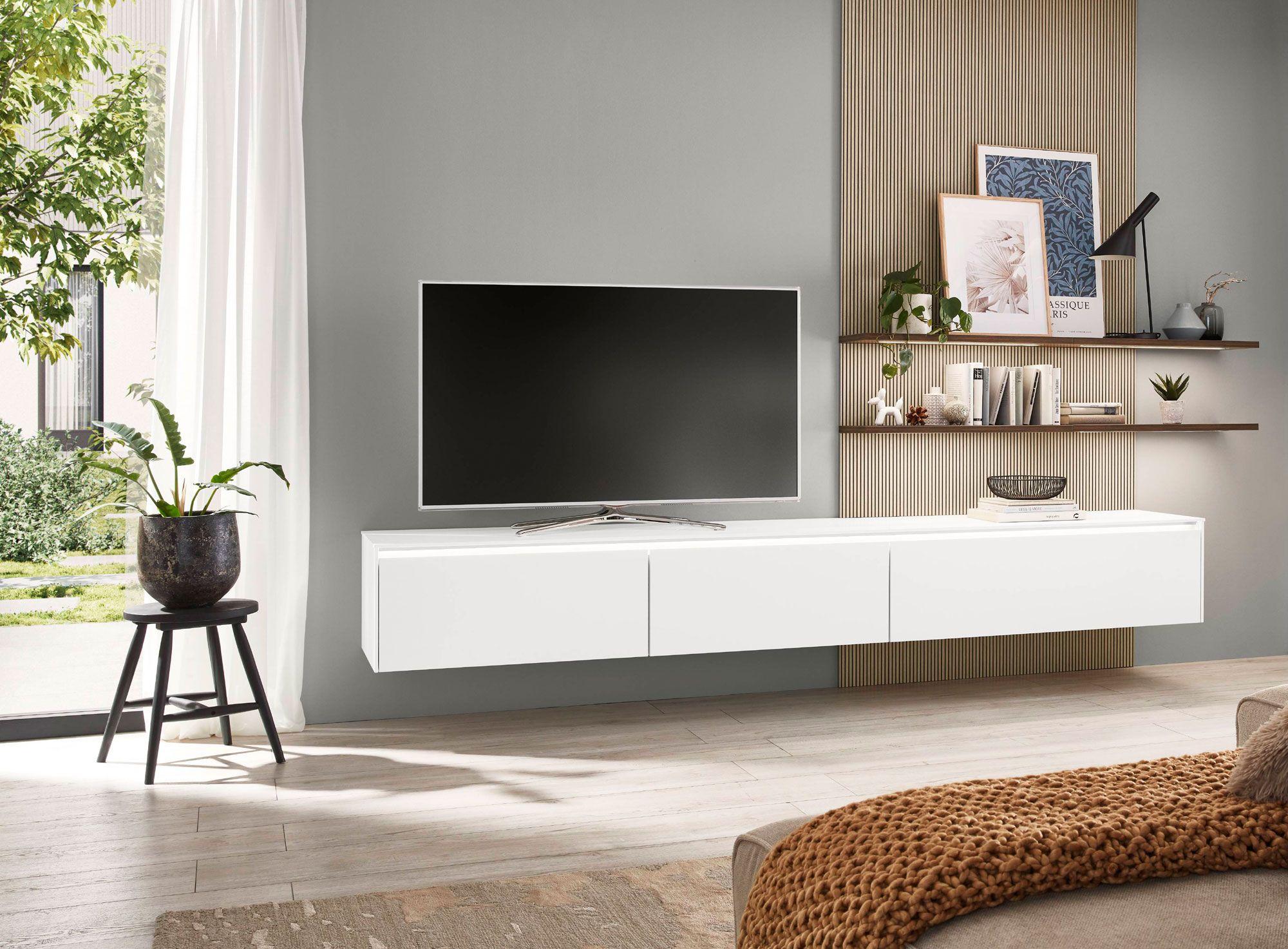 Schwebendes weißes Lowboard mit Fernseher an einer modernen Wand, ergänzt durch Holzdetails, dekorative Regale und Pflanzen, in einem lichtdurchfluteten Raum.