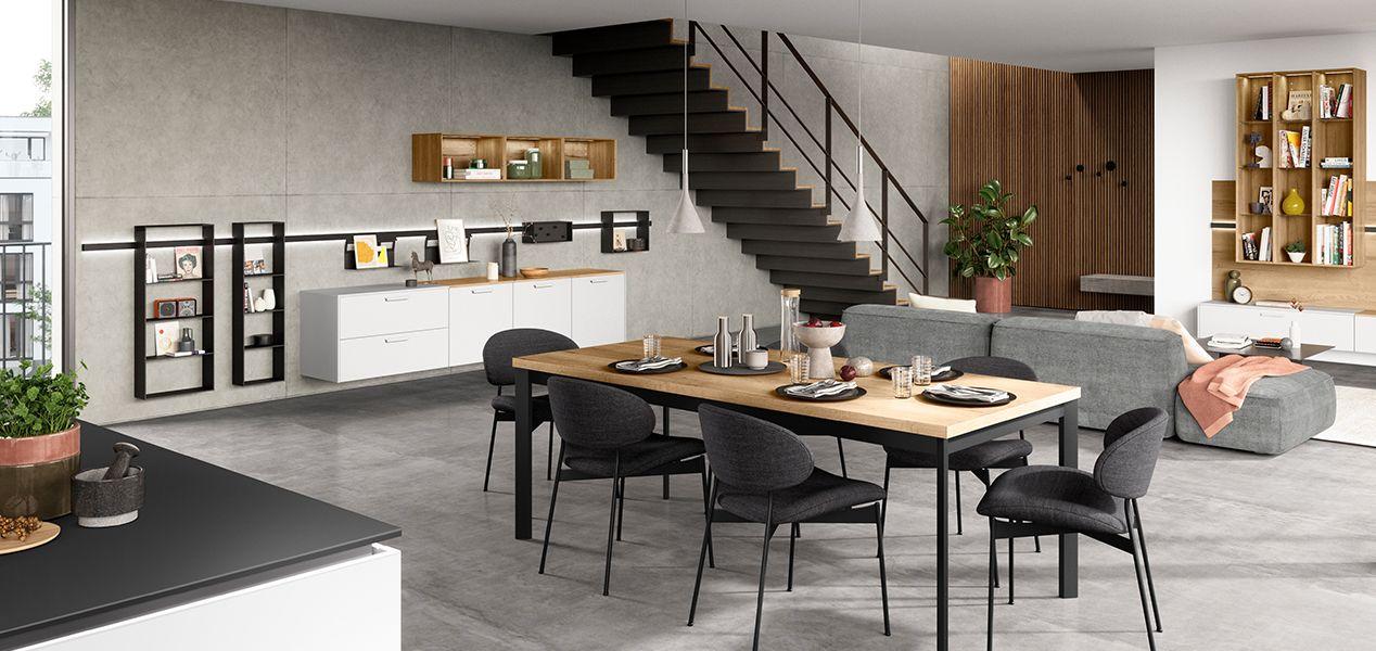 Ein modernes, offenes Wohn- und Esszimmer mit einem großen Esstisch, schwarzen Stühlen, einer eleganten Küche und einer Treppe, die zu einem oberen Bereich führt. Der Raum ist mit Pflanzen und stilvollen Regalen dekoriert.