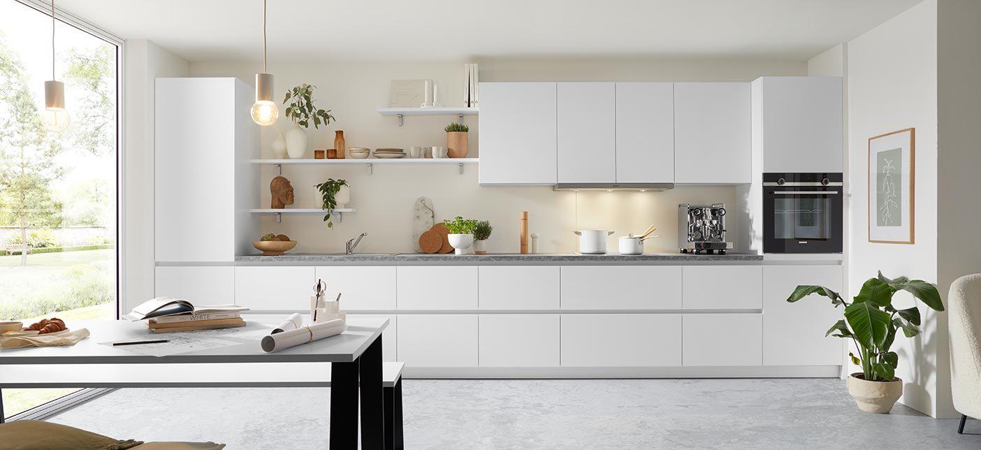 Helle, minimalistische Küche mit weißen Schränken und grauer Arbeitsplatte. Offene Regale über der Arbeitsfläche sind mit dekorativen Elementen und Geschirr bestückt. Ein großer Fensterbereich lässt viel natürliches Licht herein. Eine Kaffeemaschine und Backofen sind in die Schrankwand integriert. Pflanzen verleihen dem Raum eine frische Atmosphäre, während ein moderner Esstisch im Vordergrund zu sehen ist.