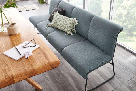 Elegante Sitzbank mit grauem Polster und schwarzem Metallgestell, dekoriert mit Kissen, kombiniert mit einem massiven Holztisch auf dunklem Holzboden und mit Blick auf große Fensterfront.