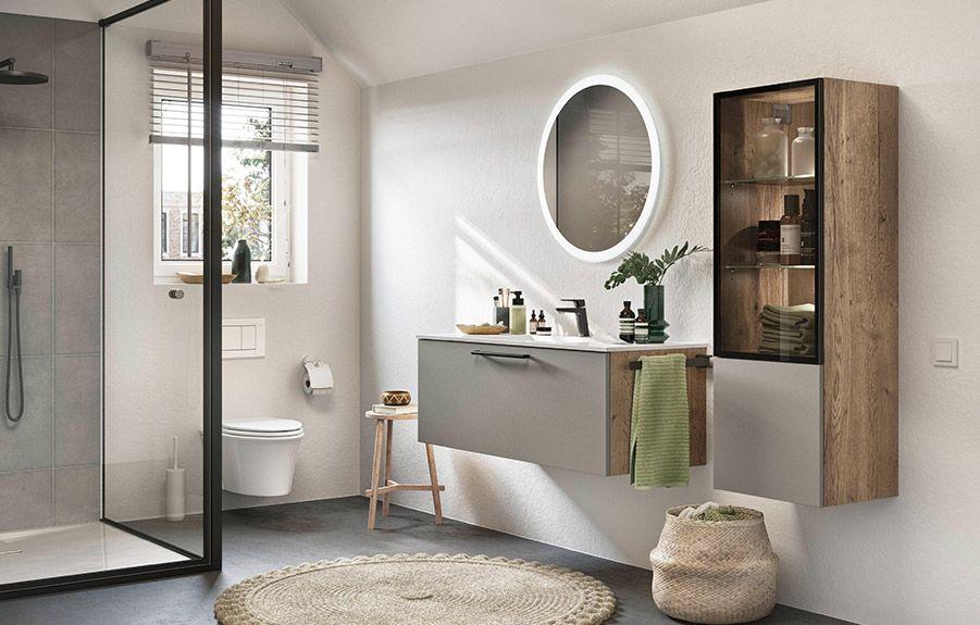Modernes Badezimmer mit offener Dusche, wandhängendem Waschtisch und rundem beleuchtetem Spiegel. Holz- und Grautöne dominieren die Gestaltung, ergänzt durch elegante Dekorationen und Pflanzen. Ein offenes Regal bietet zusätzlichen Stauraum, während der Raum durch natürliches Licht erhellt wird.