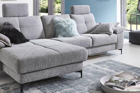 Graues Ecksofa in einem modernen Wohnzimmer mit großen Fenstern und Blick auf den Garten, dekoriert mit Kissen und einem gemütlichen Kamin im Hintergrund.