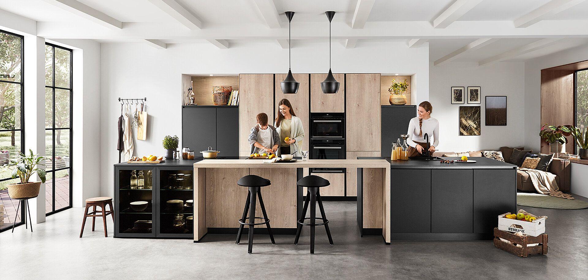 Moderne offene Wohnküche mit großer Kochinsel, Kombination aus Holz- und anthrazitfarbenen Fronten, schwarzen Pendelleuchten und Barhockern, Familie beim gemeinsamen Kochen, große Fensterfronten mit Blick ins Grüne.