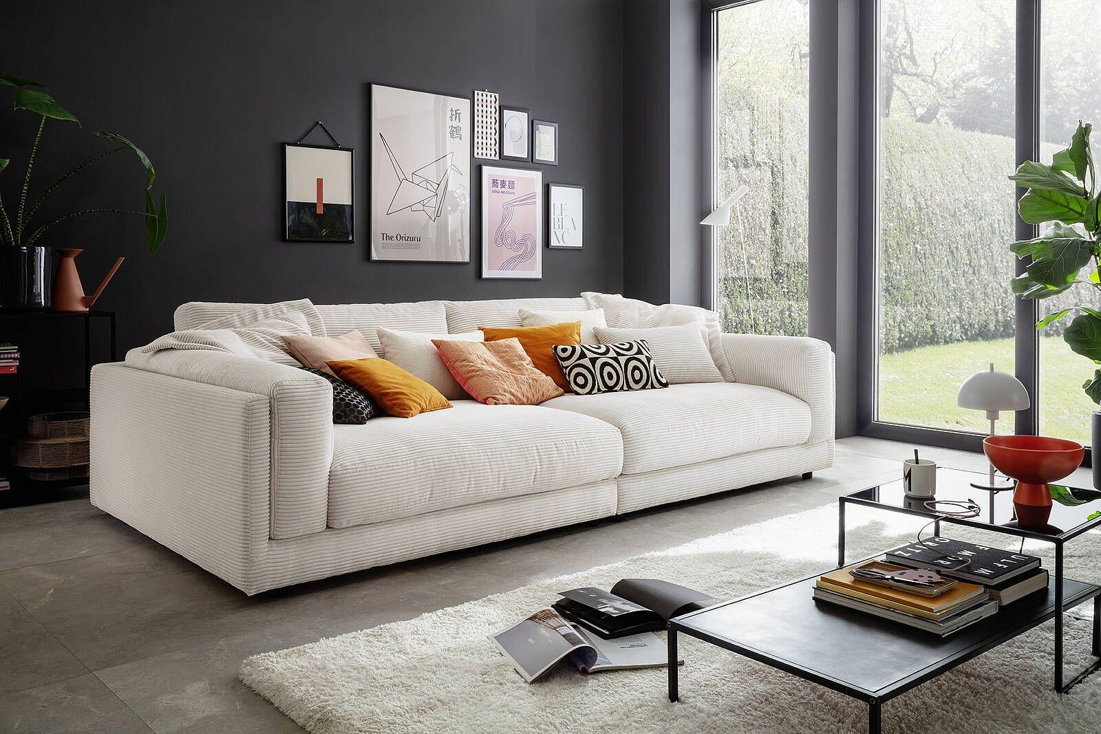 Ein stilvolles Wohnzimmer mit einem großen weißen Sofa, das mit bunten Kissen dekoriert ist, und einem niedrigen Couchtisch. An der Wand hängen verschiedene gerahmte Kunstwerke, während große Fenster einen Blick auf den Garten bieten.