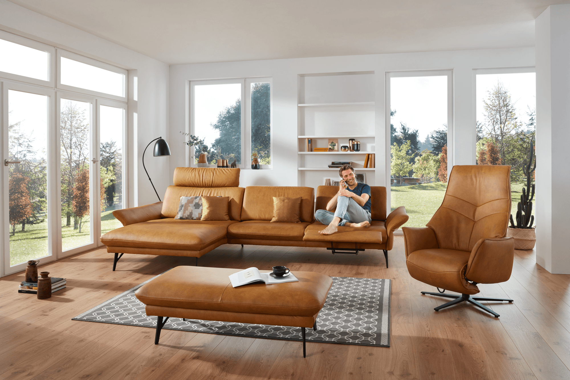 Modernes Wohnzimmer mit cognacfarbenen Ledermöbeln, Mann entspannt auf Ecksofa sitzend, große Fenster mit Gartenblick, heller Holzboden.