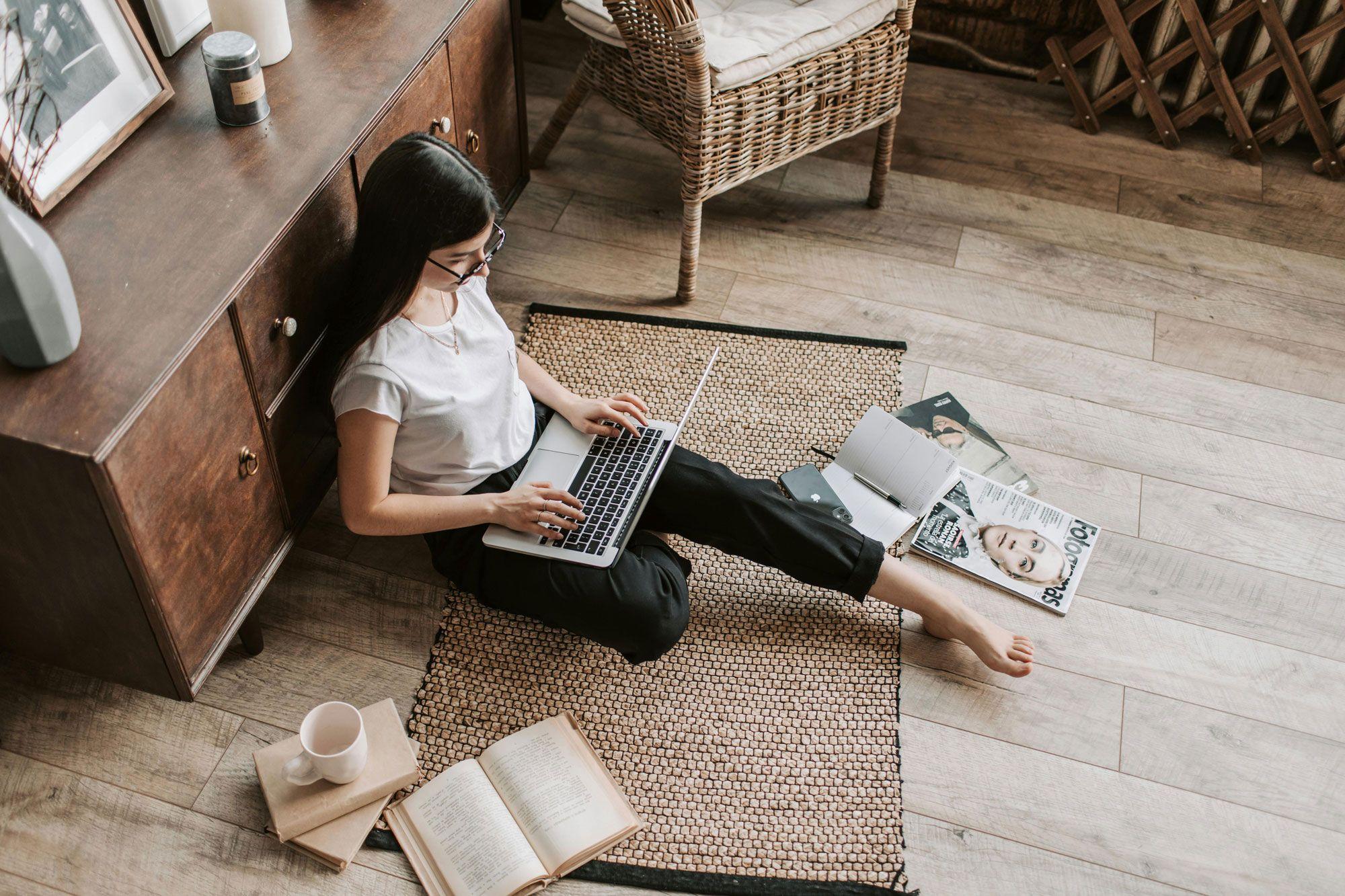 Eine Person sitzt barfuß auf einem Teppichboden, lehnt an einem Schrank und arbeitet an einem Laptop. Um sie herum liegen Bücher, eine Tasse und Zeitschriften verstreut.