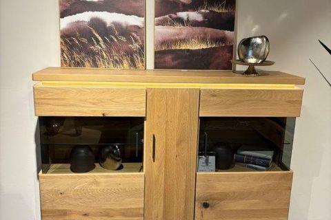 Massives Highboard aus Holz mit Glas-Elementen und vier Schubladen. Das Möbelstück ist dekoriert mit einer silbernen Vase und einer Schale auf der Oberseite. An der Wand darüber hängen zwei Kunstwerke mit Naturmotiven in warmen Farbtönen. Rechts daneben steht eine Pflanze in einem modernen, hellen Pflanzgefäß.
