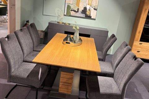 Essgruppe mit rechteckigem Holztisch auf modernem Mittelfuß in Grau mit Holzelementen. Umgeben von sechs grauen Freischwinger-Stühlen mit Polsterung. Im Hintergrund eine grüne Wand mit abstraktem Gemälde und Dekorationen.