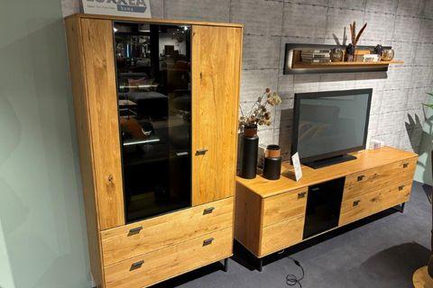 Wohnwand im rustikalen Design mit warmen Holzoberflächen. Links ein Schrank mit Glastür und Schubladen, rechts ein TV-Board mit Fernseher und dekorativer Rückwand. Darüber ein Wandregal mit Dekorationselementen.