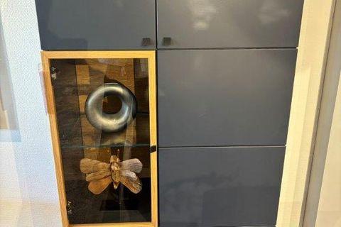 Dunkelgraue Vitrine mit Holzakzenten und einer beleuchteten Glasvitrine, die mit Gläsern dekoriert ist. Oberhalb eine große Schmetterlingsdekoration aus Holz. Steht vor einer schlichten Wand auf filigranen Metallfüßen.