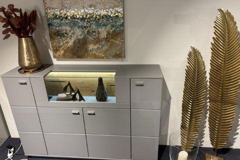 Graues Highboard mit zentralem beleuchtetem Fach, dekoriert mit Skulpturen und Vasen. Auf der Oberseite eine große goldene Vase mit roten Zweigen. Im Hintergrund ein abstraktes Gemälde mit Naturmotiven. Daneben goldene Deko-Elemente in Form von Blättern.