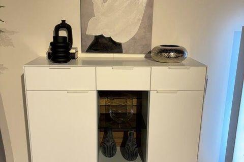 Weißes Highboard mit zentraler Glasvitrine und modernen Dekorationen, darunter Vasen und eine Schale. Daneben ein hoher grauer Pflanzentopf mit grünem Baum. Hintergrund mit heller Wand und abstraktem Kunstwerk.