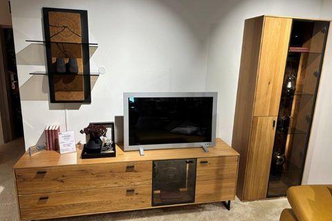 Wohnwand mit TV-Board aus hellem Holz, darauf ein Fernseher und Dekorationen. Rechts ein hohes Vitrinenschrank mit Glastüren und Beleuchtung, gefüllt mit Dekorationsgegenständen. Links ein modernes Wandregal mit schwarzem Rahmen und Holzdetails.