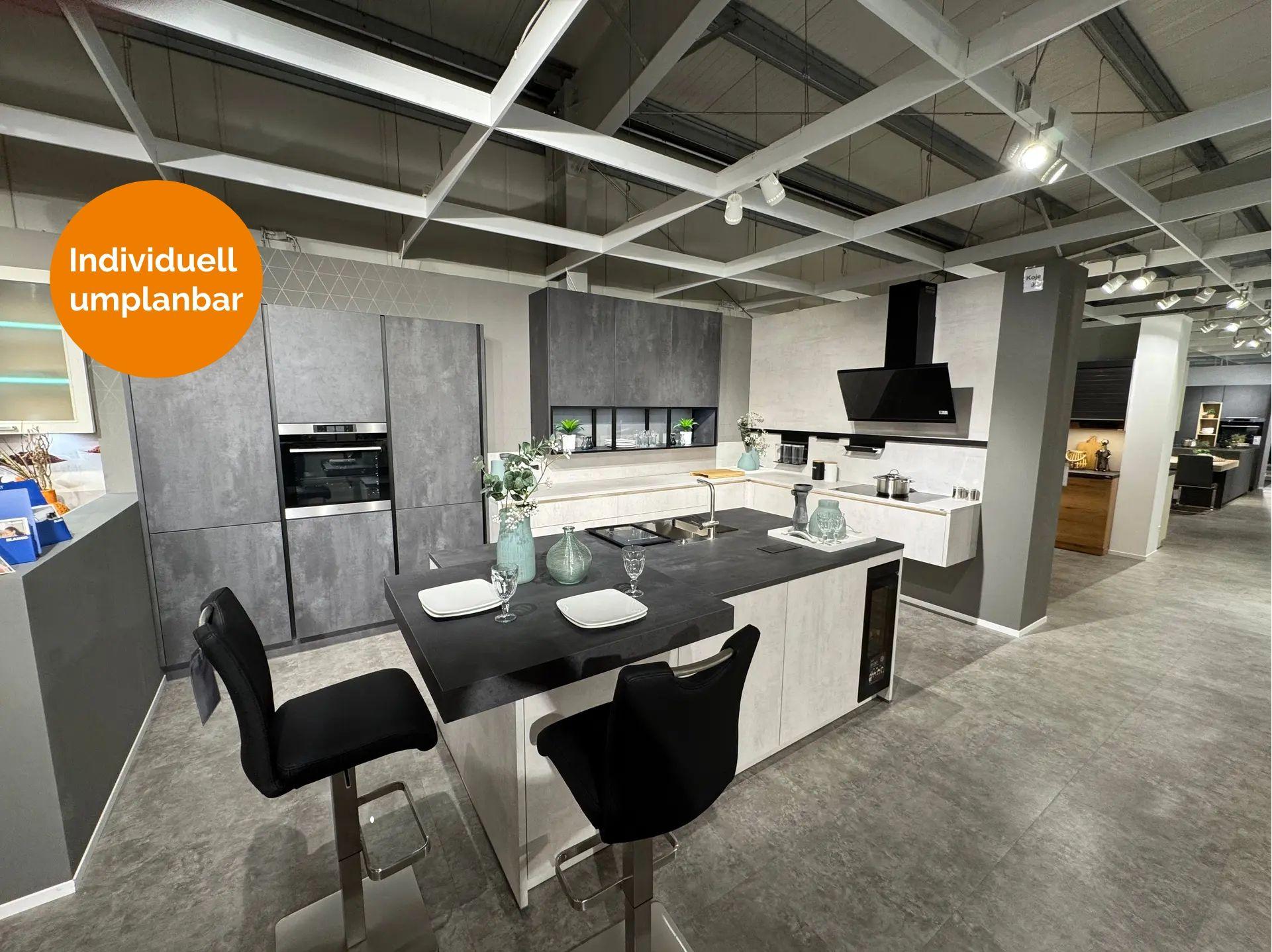 Zweifarbige Küche in heller und dunkler Betonoptik mit großer Kochinsel, schwarzem Bartresen, Barhockern, integriertem Weinkühlschrank, Glasvitrine und schwarzer Schräghaube.