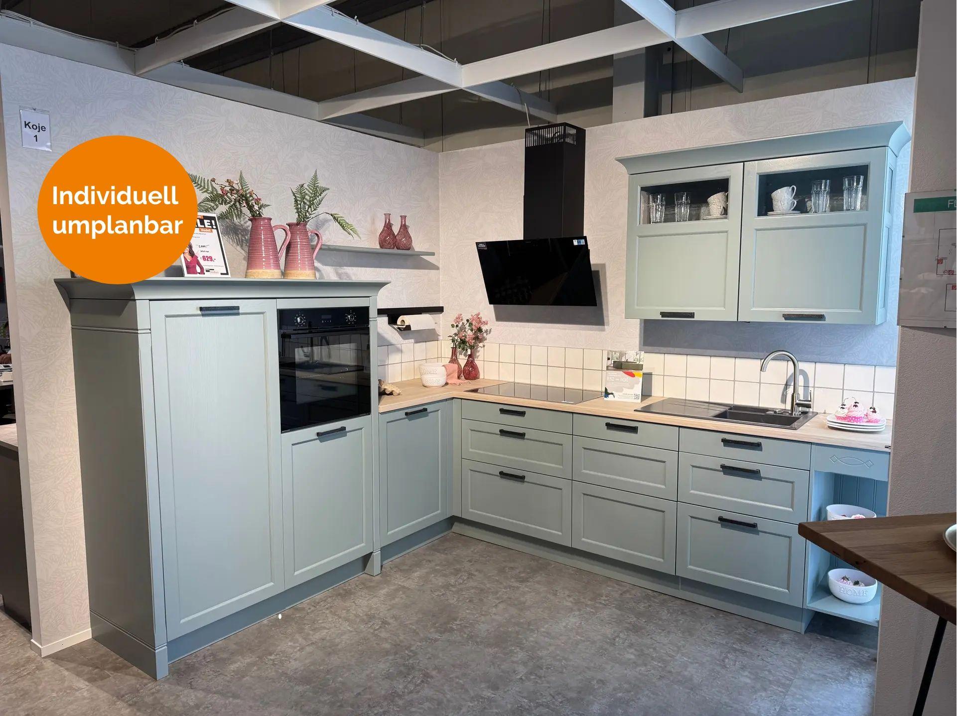 Landhausküche in Salbeigrün mit Rahmenfronten, schwarzen Griffen, Holzarbeitsplatte und schwarzer Dunstabzugshaube in L-Form – Ausstellung Koje 1.