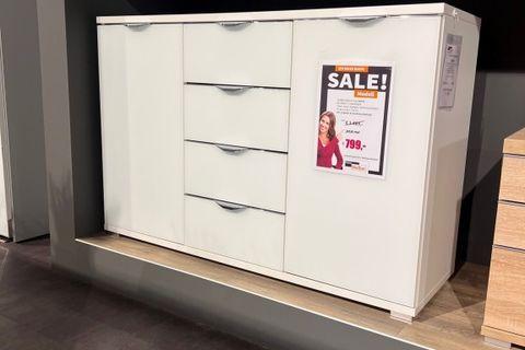 Weiße Großraumkommode mit drei Schubladen in der Mitte und Türen an den Seiten. Präsentiert auf einem Podest mit einem SALE-Schild, das einen Sonderpreis von 799€ anzeigt. Steht in einer modernen Ausstellung.