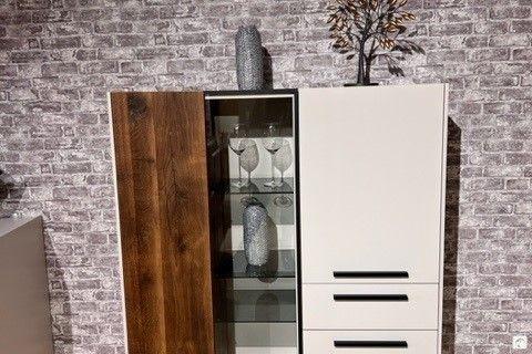 Modernes Highboard mit einer Kombination aus dunklen Holz- und weißen Fronten, einer zentralen Glasvitrine mit dekorativen Gläsern und Vasen. Steht vor einer grauen Backsteinwand und ist mit weiteren Deko-Elementen auf der Oberseite verziert.