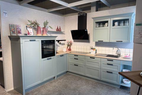 Landhausküche in Salbeigrün mit Rahmenfronten, schwarzen Griffen, Holzarbeitsplatte und schwarzer Dunstabzugshaube in L-Form – Ausstellung Koje 1.