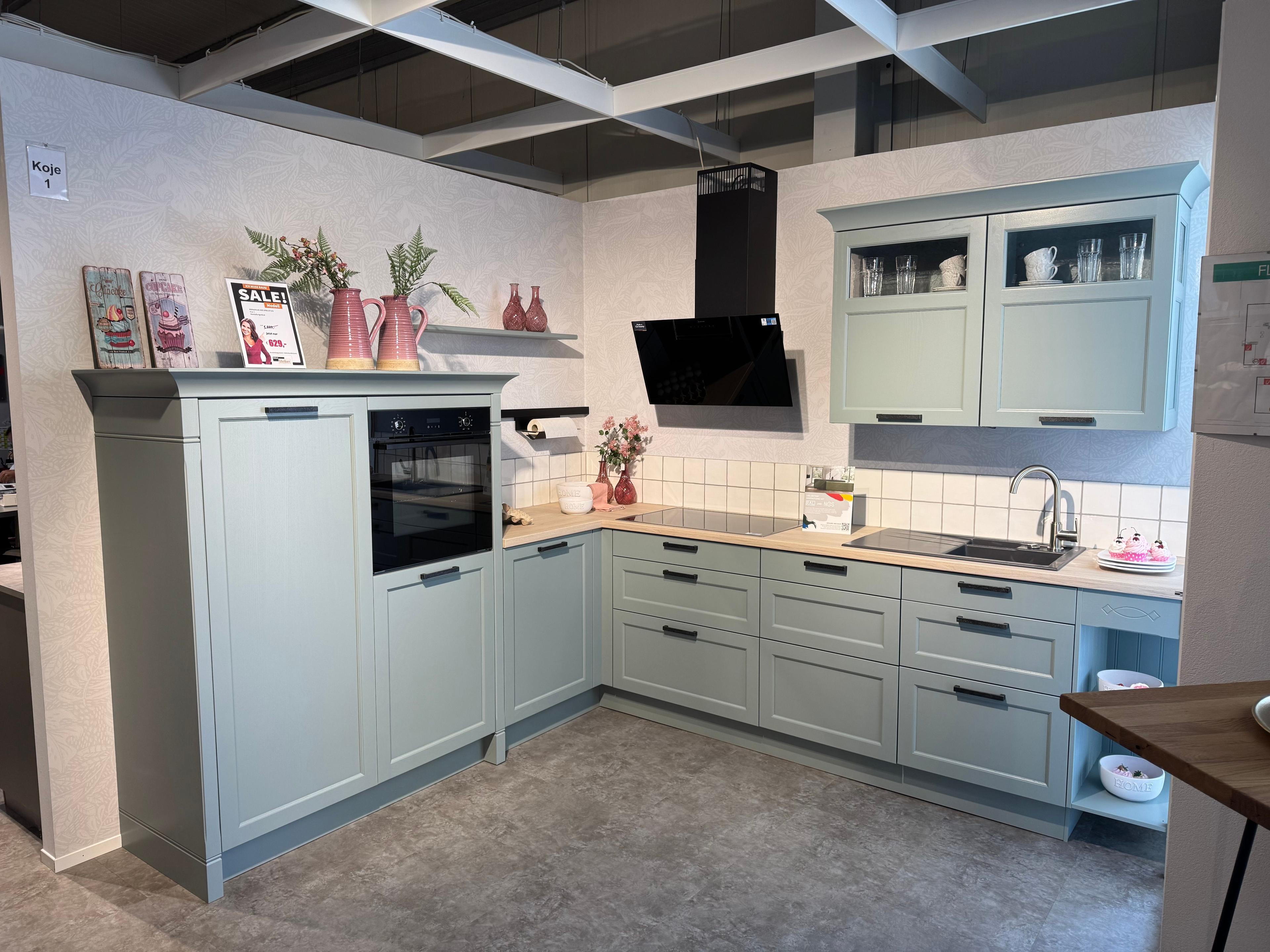 Landhausküche in Salbeigrün mit Rahmenfronten, schwarzen Griffen, Holzarbeitsplatte und schwarzer Dunstabzugshaube in L-Form – Ausstellung Koje 1.