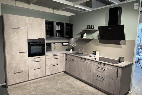 Moderne Küche in Betonoptik mit schwarzen Griffen, Glasvitrine, schwarzer Schräghaube und grauer Arbeitsplatte in L-Form – Ausstellung Koje 2.