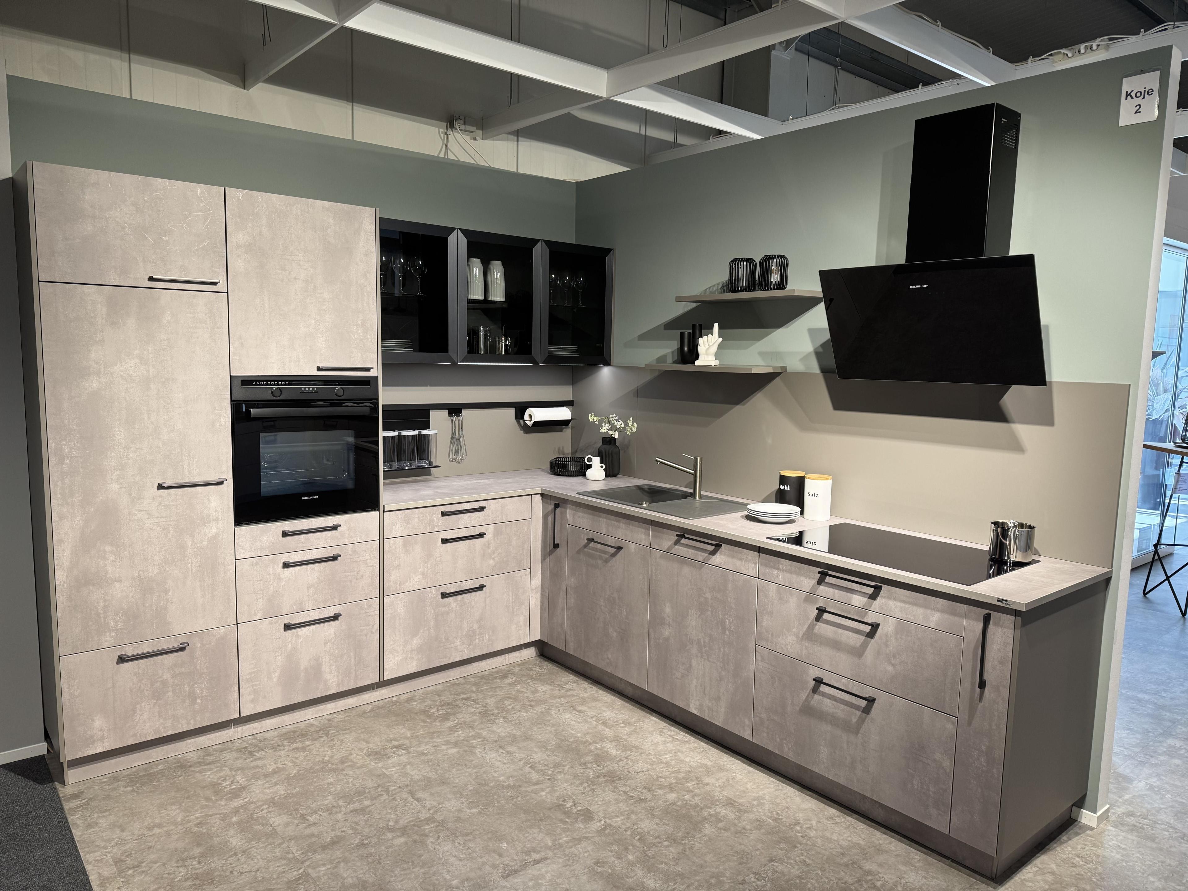 Moderne Küche in Betonoptik mit schwarzen Griffen, Glasvitrine, schwarzer Schräghaube und grauer Arbeitsplatte in L-Form – Ausstellung Koje 2.