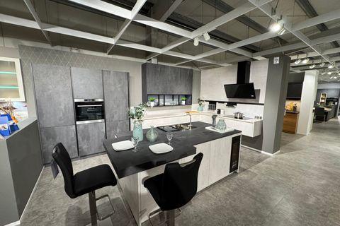 Zweifarbige Küche in heller und dunkler Betonoptik mit großer Kochinsel, schwarzem Bartresen, Barhockern, integriertem Weinkühlschrank, Glasvitrine und schwarzer Schräghaube.