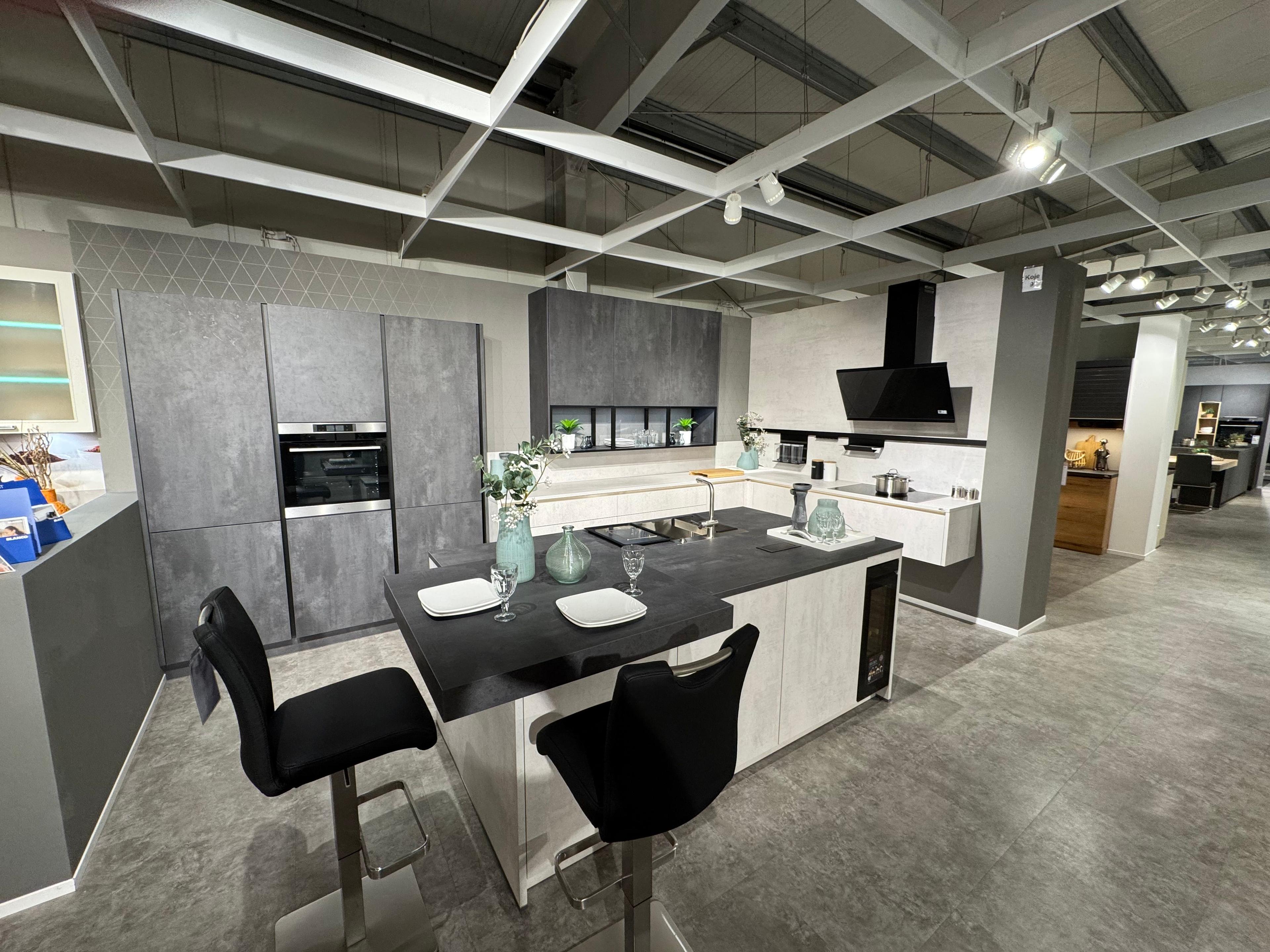 Zweifarbige Küche in heller und dunkler Betonoptik mit großer Kochinsel, schwarzem Bartresen, Barhockern, integriertem Weinkühlschrank, Glasvitrine und schwarzer Schräghaube.