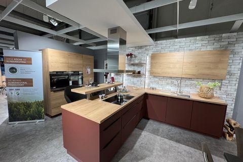 Klimaneutrale Küche in Terrakottarot und Eiche-Optik mit Kochinsel, integriertem Essplatz, Edelstahl-Inselhaube, Steinwand-Verblender und Hochschrank mit Einbaugeräten.