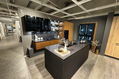 Elegante Küche in Mattschwarz und Nussbaum-Optik mit freistehender Kochinsel, beleuchteten Glasvitrinen, offenen Designregalen und integriertem Geräteschrank.