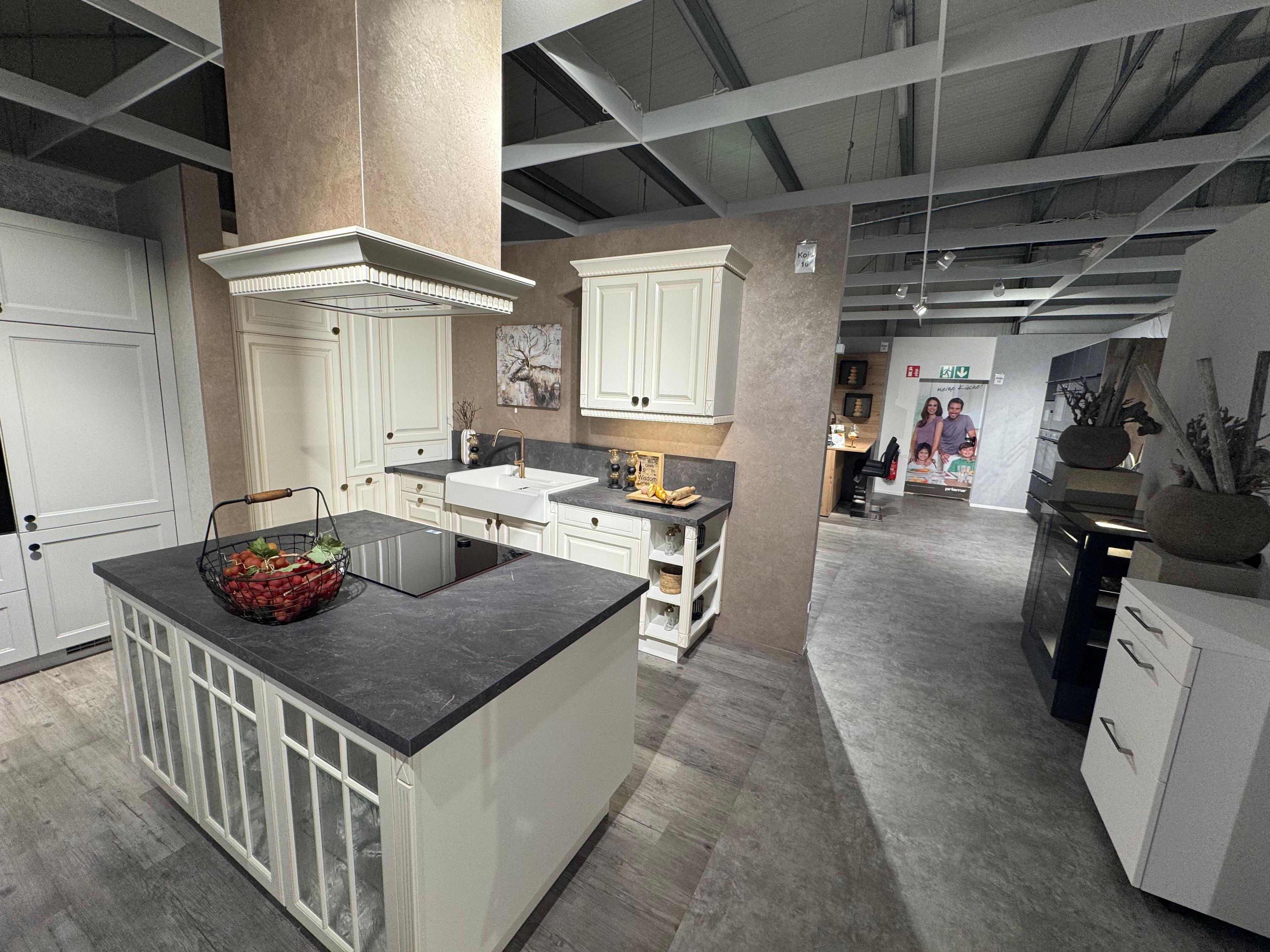 Klassische Landhausküche in Cremeweiß mit Kassettenfronten, dunkler Steinarbeitsplatte, Kochinsel mit Sprossenverglasung, Keramik-Spülbecken, Messingarmatur und dekorativer Haubenverkleidung im Landhausstil.