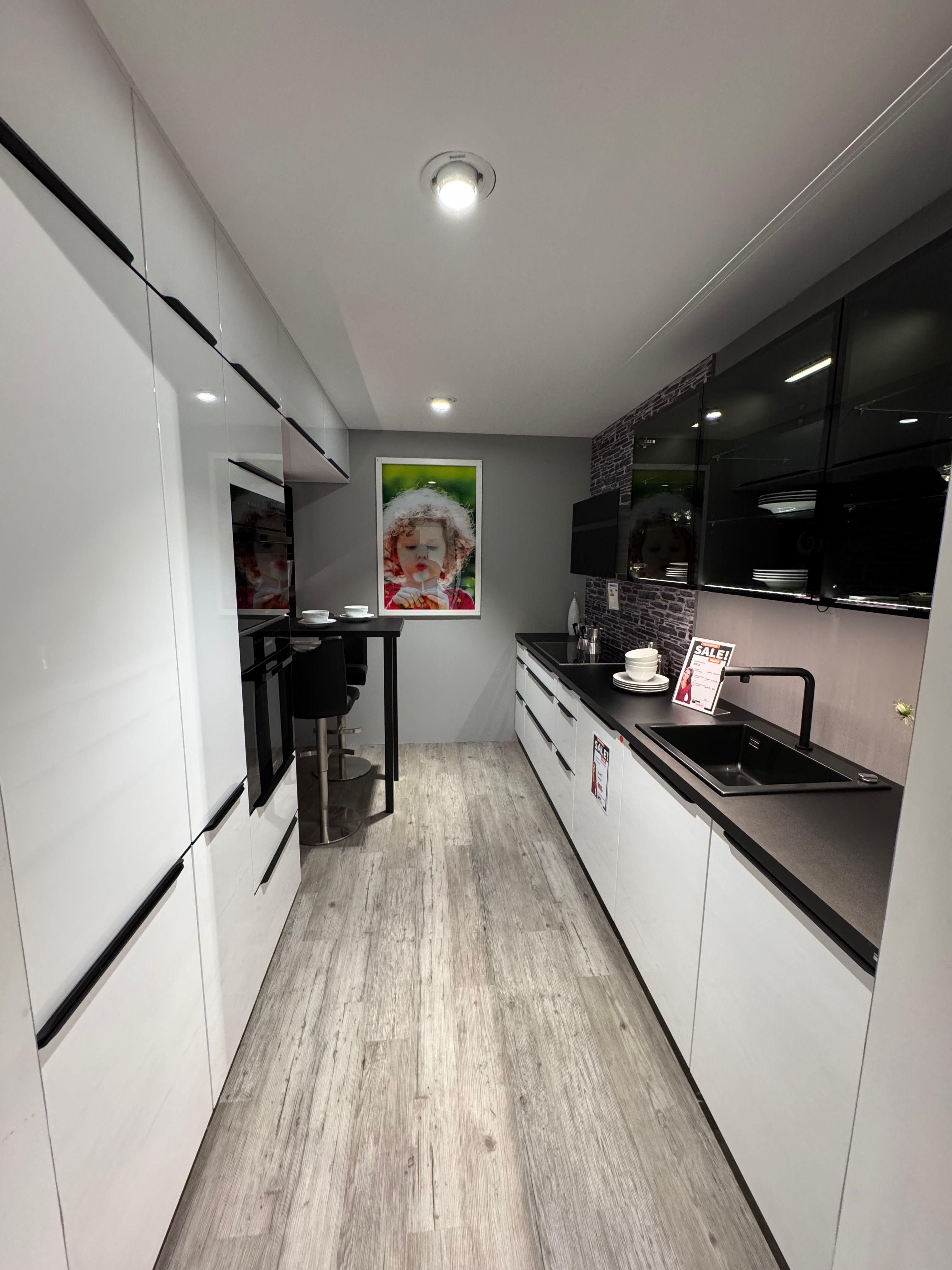 Küche in Weiß Hochglanz mit schwarzen Griffleisten, dunkler Arbeitsplatte, schwarzen Glas-Hängeschränken mit Innenbeleuchtung, Steinoptik-Nischenrückwand, schwarzer Armatur und kleinem Bartisch mit Hockern