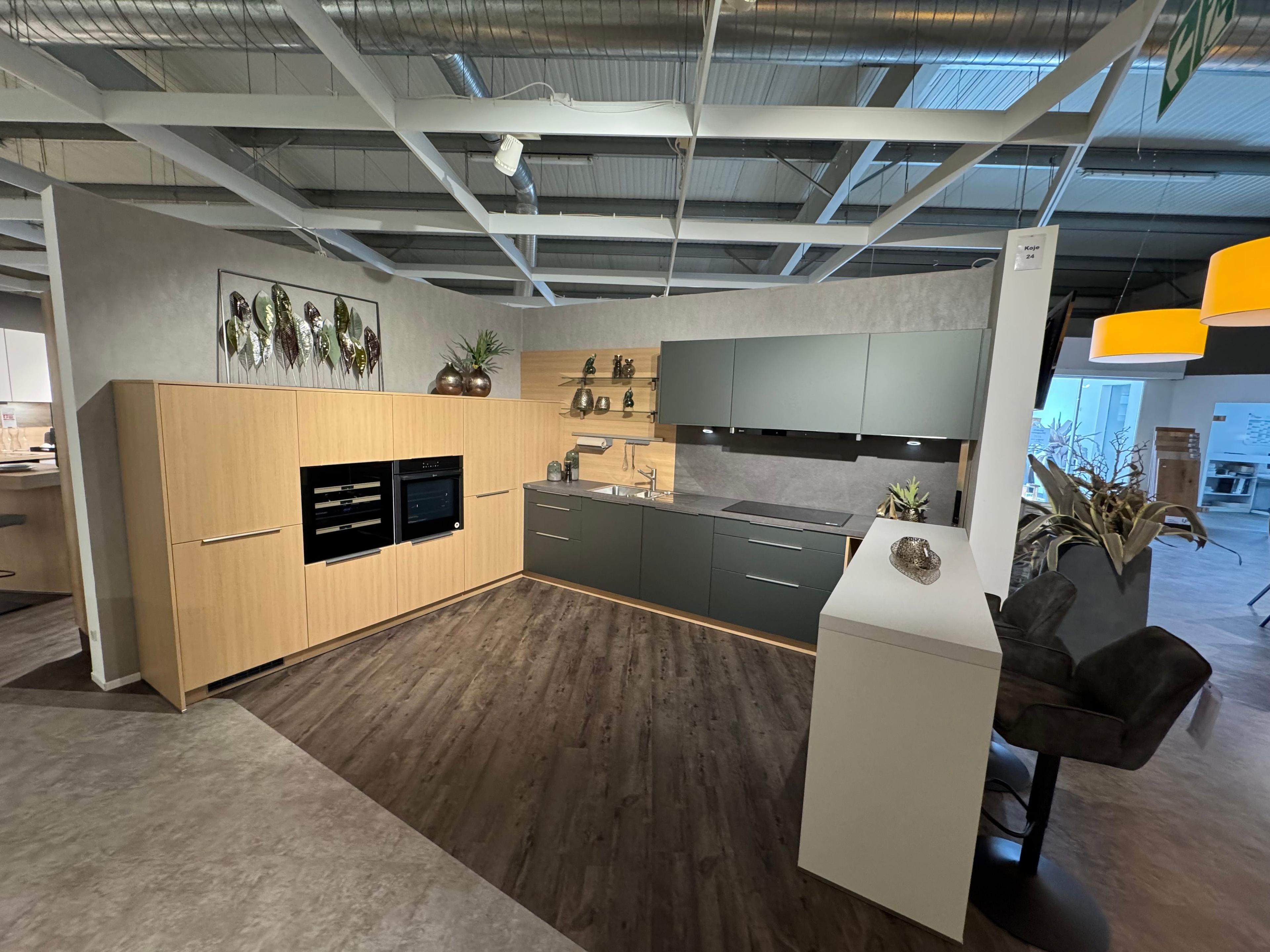 L-Küche in Metallic Ggrün und heller Eiche-Optik mit dunkler Arbeitsplatte, Holz-Nischenrückwand mit Relingsystem, Hochschrank mit Einbaugeräten, weißem Bartresen mit braunen Polsterhockern und dekorativer Blätter-Wanddeko.