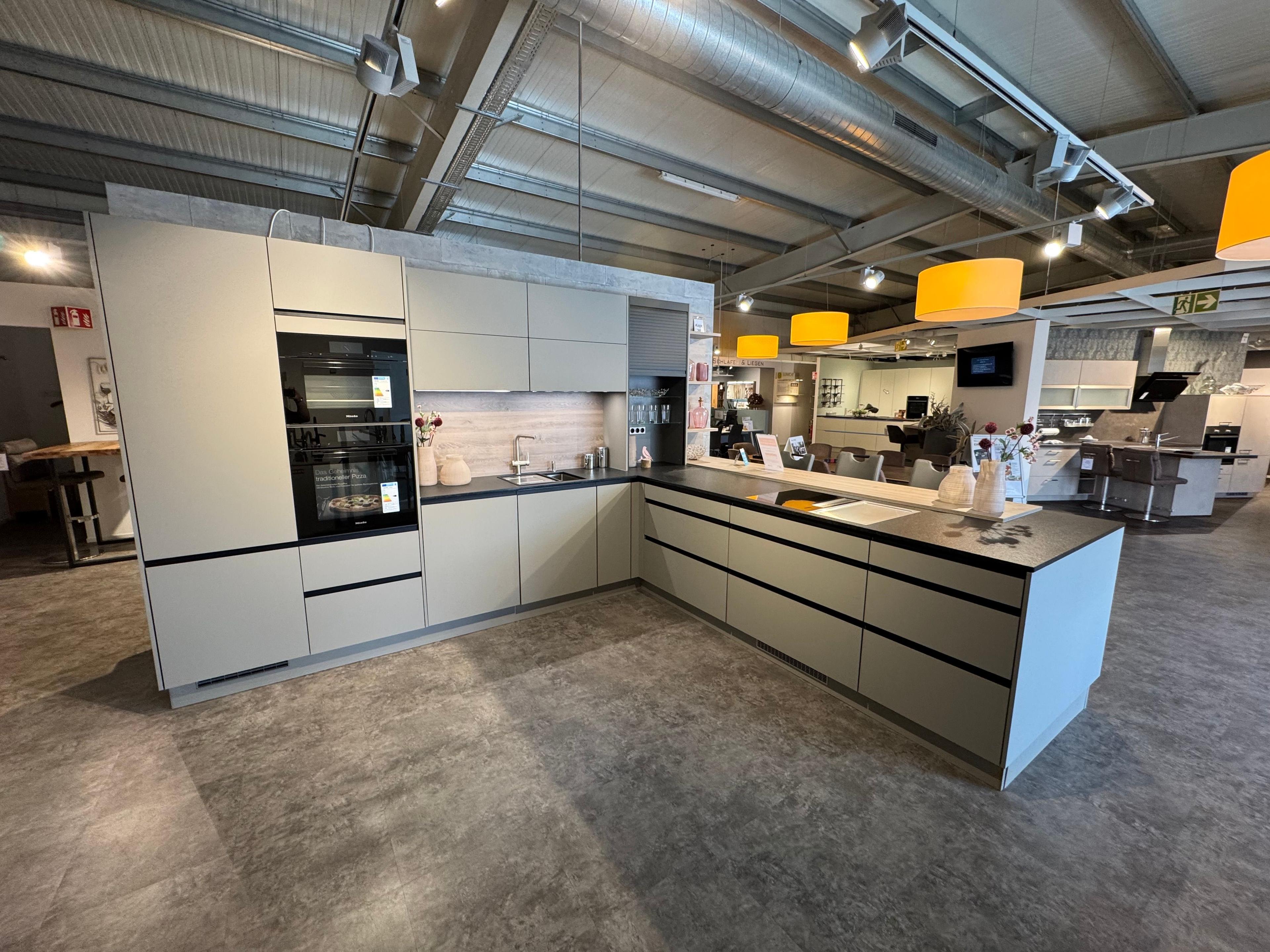 Großzügige U-Küche in mattem Graubeige mit schwarzen Griffmulden, dunkler Arbeitsplatte, Holz-Nischenrückwand, Hochschrank mit Miele-Einbaugeräten und Halbinsel mit Kochfeld.