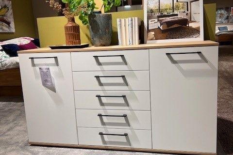Weiße Kommode mit sechs zentralen Schubladen und zwei Türen an den Seiten, ergänzt durch eine Holzoberfläche. Dekoriert mit grünen Pflanzen in Vasen und einem Werbeaufsteller für „Casada Calvia“. Steht in einer modernen Wohnraum-Ausstellung.