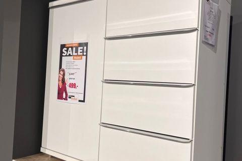 Weiße Kommode mit drei großen Schubladen und einer Tür auf der linken Seite. Präsentiert auf einem Podest mit einem SALE-Schild, das einen Sonderpreis von 499€ zeigt. Moderner und minimalistischer Stil in einer Ausstellung.