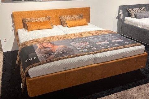 Polsterbett in warmem Orangeton mit hohem Kopfteil und passender Bettwäsche in Braun- und Beigetönen. Auf dem Bett eine Werbedecke mit Produktinformationen. Hintergrund mit Spiegelwand und weiteren Betten in einer Ausstellung.