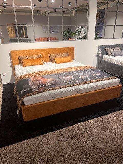 Polsterbett in warmem Orangeton mit hohem Kopfteil und passender Bettwäsche in Braun- und Beigetönen. Auf dem Bett eine Werbedecke mit Produktinformationen. Hintergrund mit Spiegelwand und weiteren Betten in einer Ausstellung.