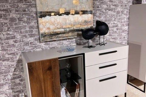 Modernes Sideboard mit weißen Fronten, dunklen Holzelementen und einer zentralen Glasvitrine, dekoriert mit Büchern und Vasen. Auf der Oberseite schwarze Skulpturen und Dekorationen. Hintergrund mit grauer Backsteinwand und abstraktem Gemälde in neutralen Farben.