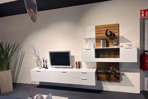 Wohnwand im minimalistischen Stil mit weißem, schwebendem TV-Board, einem kleinen Fernseher und Dekoration. Rechts ein Regal mit weißen und holzfarbenen Elementen, dekoriert mit Büchern, Gläsern und Skulpturen. Im Hintergrund eine helle Wand mit Pflanzen und Vasen.
