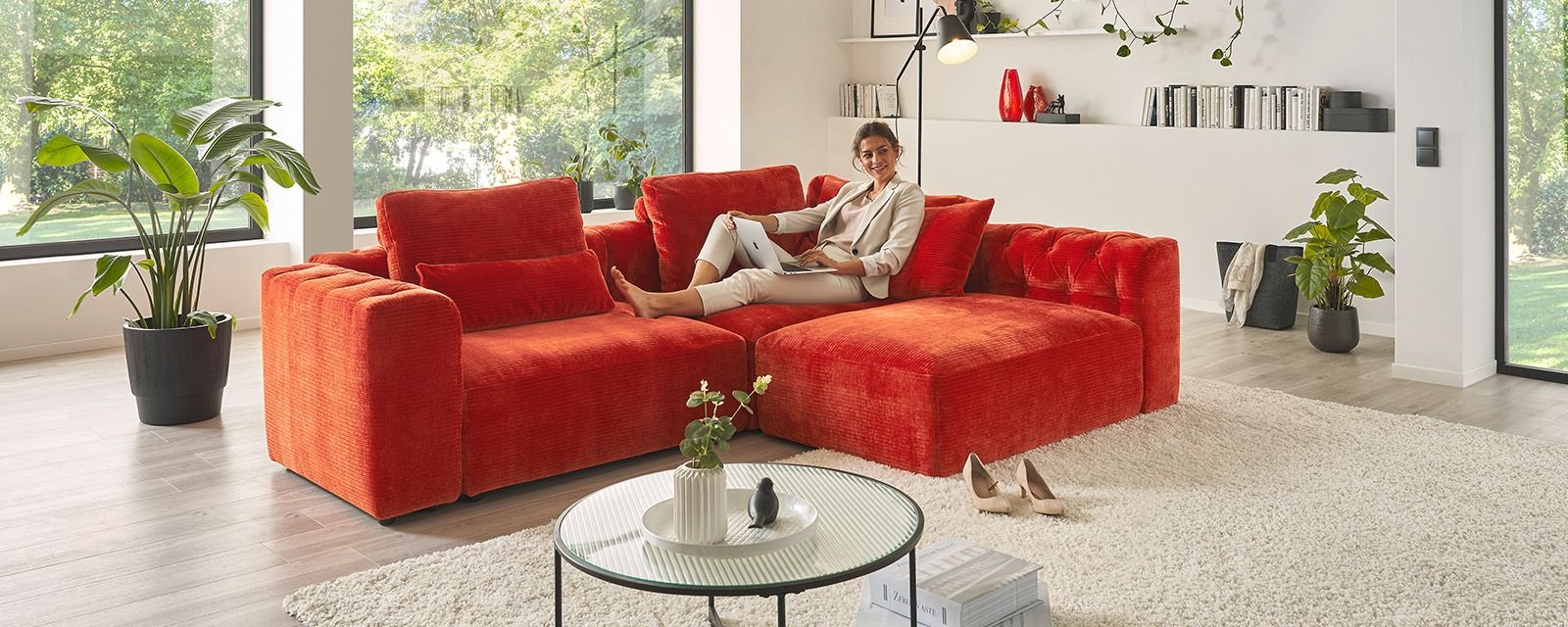 Rotes Sofa in modernem Wohnzimmer mit großen Fenstern, Pflanzen und dekorativen Elementen, auf dem eine Frau mit Laptop sitzt.