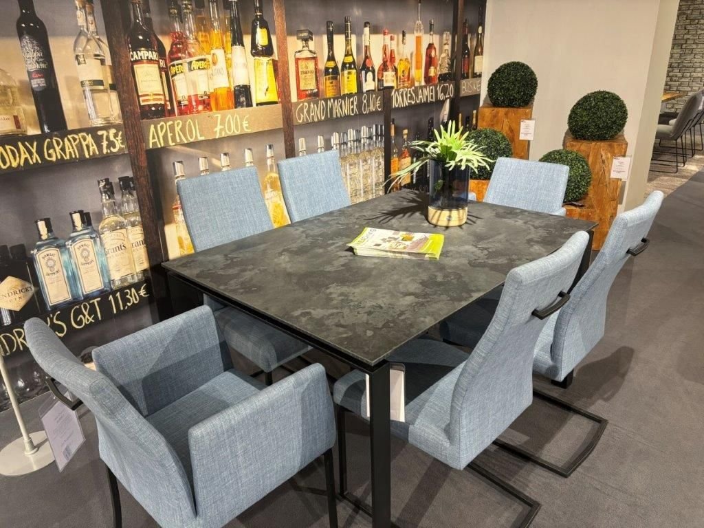 Essgruppe mit quadratischem Tisch und dunkler Steinoptik-Tischplatte, umgeben von sechs hellblauen Polsterstühlen, teils mit Armlehnen und Freischwinger-Gestell. Hintergrund mit dekorativer Bar-Wand und grünen Buchsbaum-Elementen.