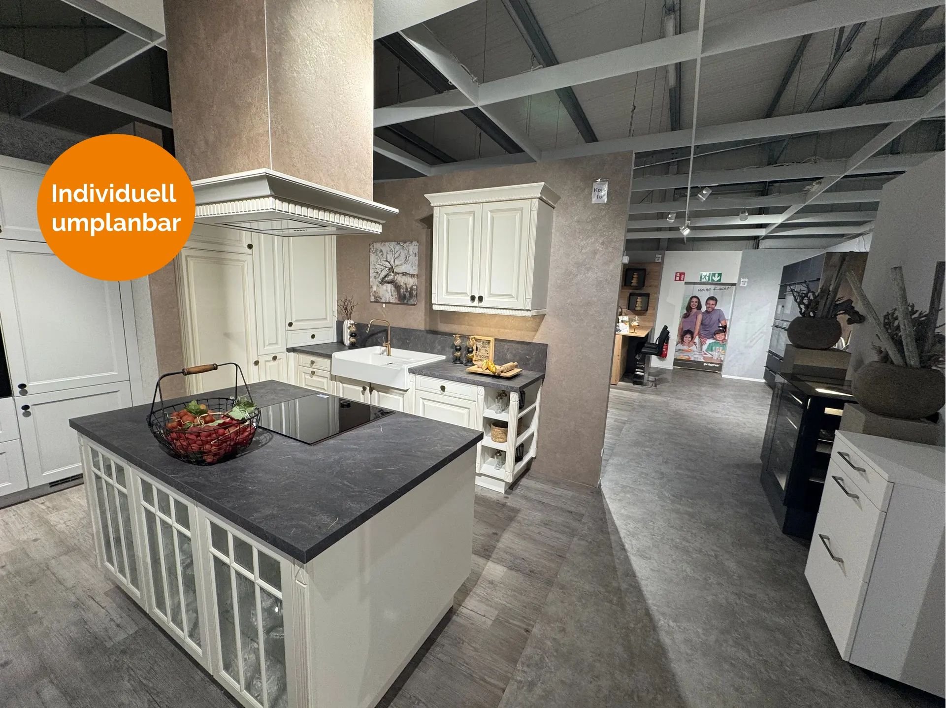 Klassische Landhausküche in Cremeweiß mit Kassettenfronten, dunkler Steinarbeitsplatte, Kochinsel mit Sprossenverglasung, Keramik-Spülbecken, Messingarmatur und dekorativer Haubenverkleidung im Landhausstil.