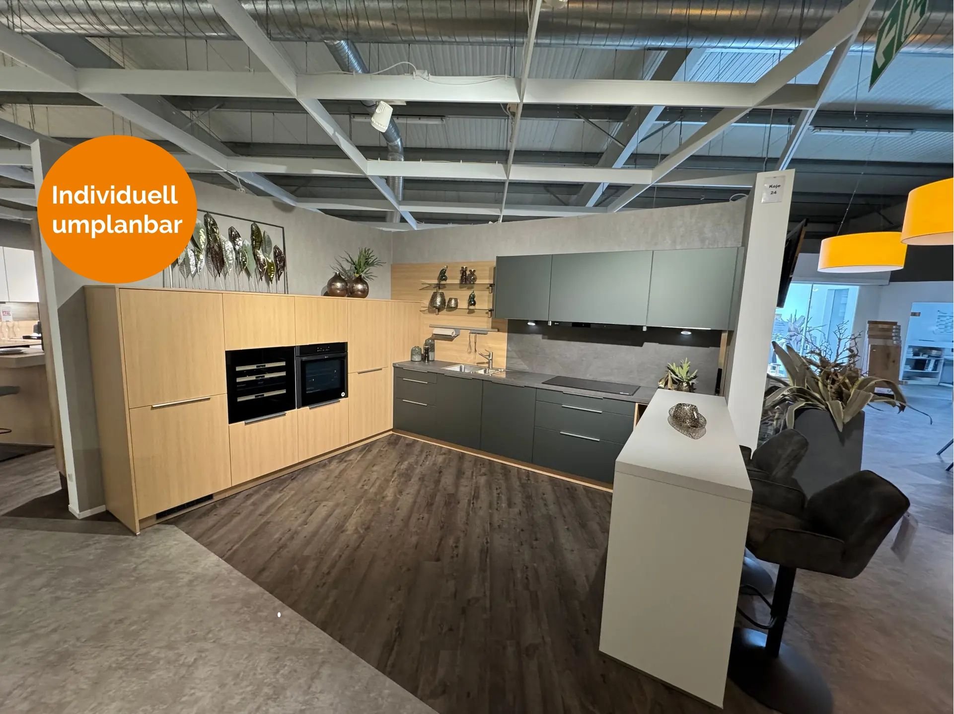 L-Küche in Metallic Ggrün und heller Eiche-Optik mit dunkler Arbeitsplatte, Holz-Nischenrückwand mit Relingsystem, Hochschrank mit Einbaugeräten, weißem Bartresen mit braunen Polsterhockern und dekorativer Blätter-Wanddeko.