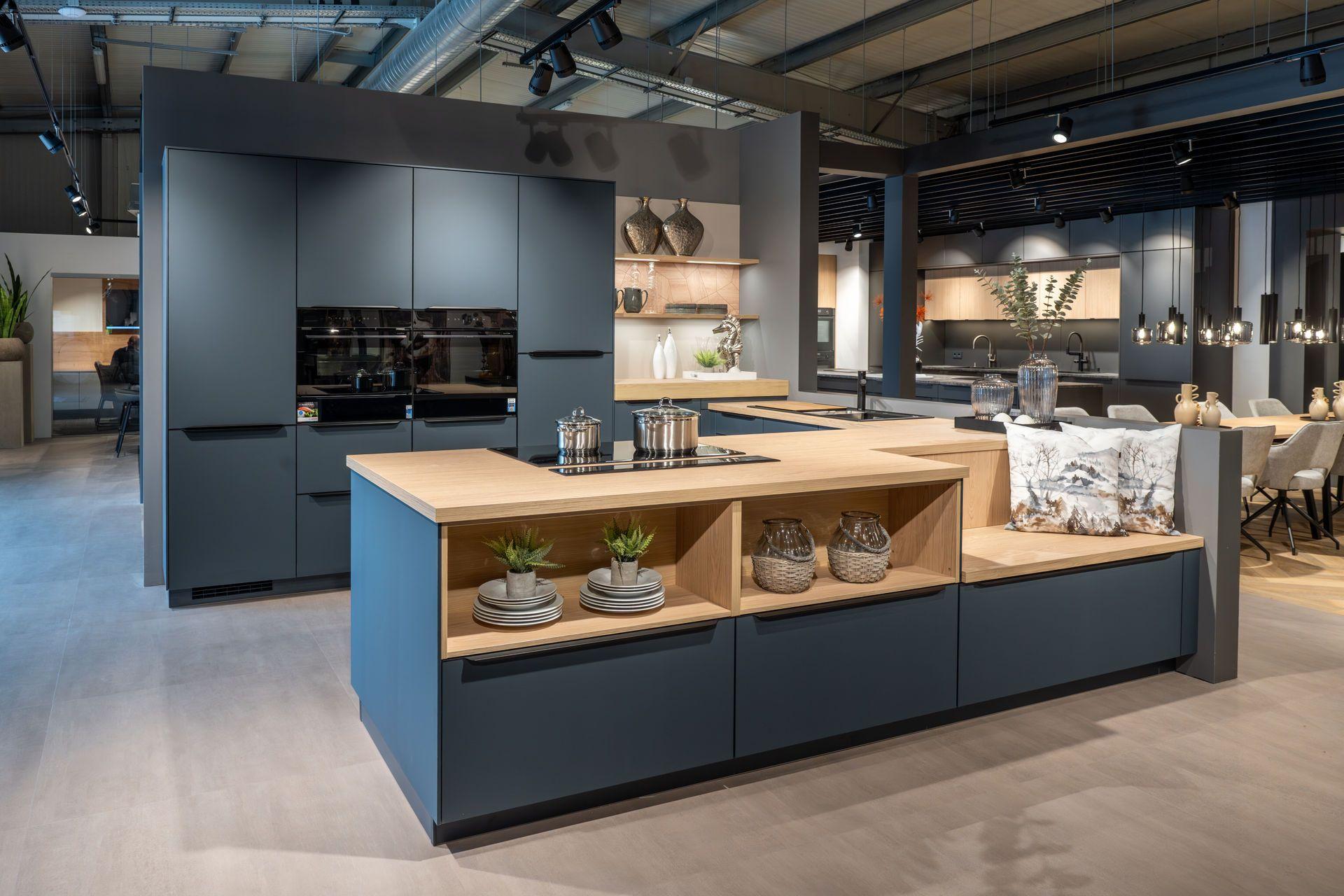 Ausstellungsküche in Blaugrau mit Holz-Arbeitsplatte, großer Kochinsel mit offenen Regalfächern, Hochschränken mit Einbaugeräten und angrenzendem Essplatz im Küchenstudio von Möbel Melber.